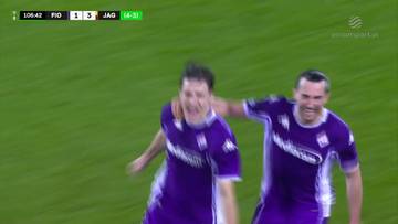 Dwóch piłkarzy w fioletowych strojach Fiorentiny cieszy się z zdobytej bramki, jeden z nich obejmuje drugiego ramieniem. Na zielonym tle murawy widoczny jest fragment tablicy wyników meczu.