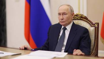 Władimir Putin siedzi przy stole, na kt&oacute;rym leżą dokumenty, a za nim widoczna jest flaga Rosji.