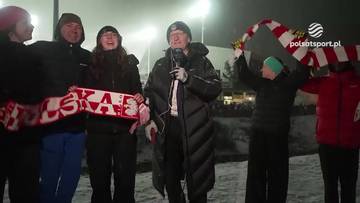 Grupa osób świętuje, trzymając szaliki z napisem POLSKA. W środku stoi mężczyzna z mikrofonem.