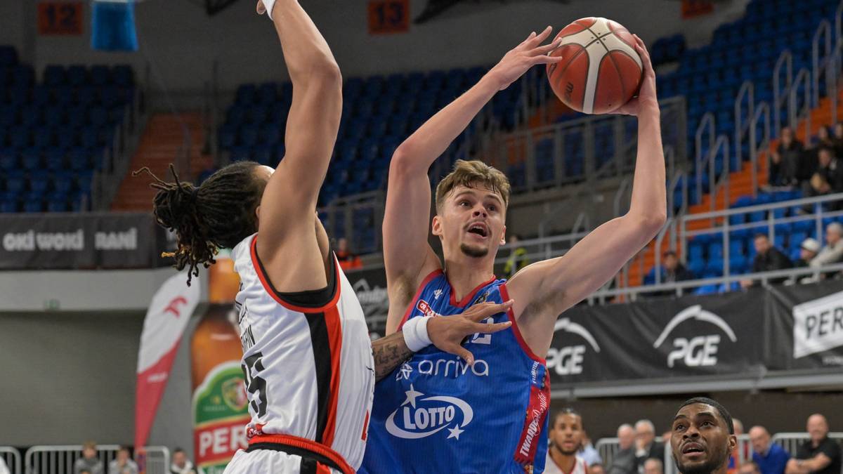 Dwóch koszykarzy walczy o piłkę podczas meczu. Zawodnik w niebieskim stroju z logo "Lotto" wykonuje rzut, podczas gdy zawodnik w białym stroju próbuje go zablokować.