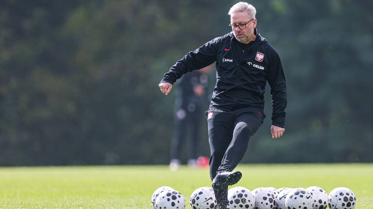 Trener w okularach, ubrany w czarną bluzę z orłem, kopie piłkę nożną na boisku.