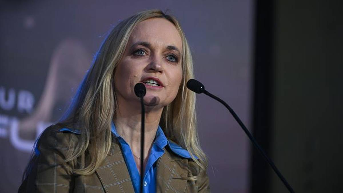 Kobieta z blond włosami, ubrana w brązowy garnitur i niebieską koszulę, przemawia do mikrofonu na scenie.