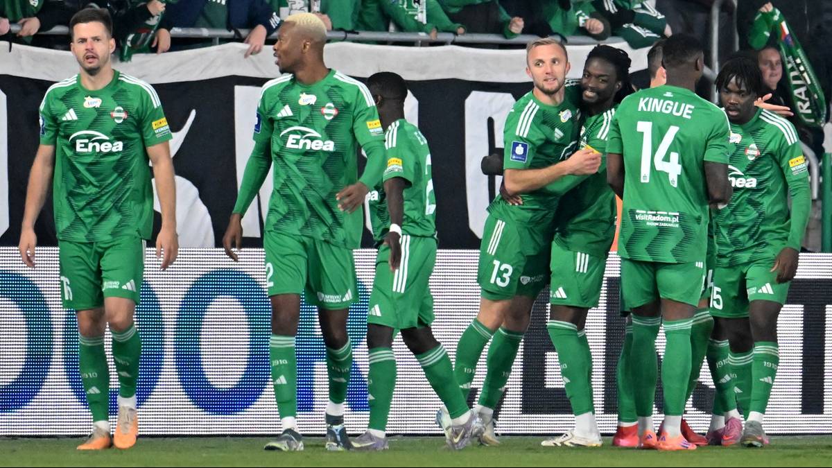 Siedmiu piłkarzy w zielonych strojach, prawdopodobnie drużyna Radomiaka, pozuje do zdjęcia na boisku piłkarskim. Widoczni są gracze z numerami 13 i 14 na koszulkach.