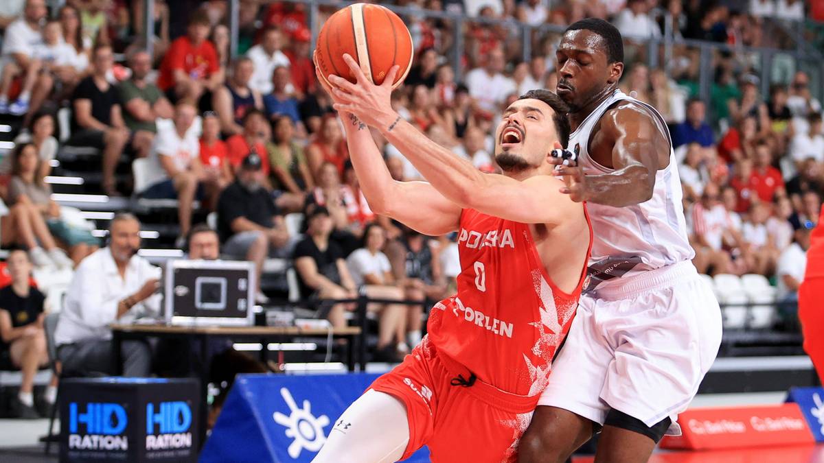 Dwaj zawodnicy koszyk&oacute;wki podczas meczu, jeden z nich w czerwonym stroju pr&oacute;buje rzucić piłkę do kosza, drugi w białym stroju stara się go zablokować.