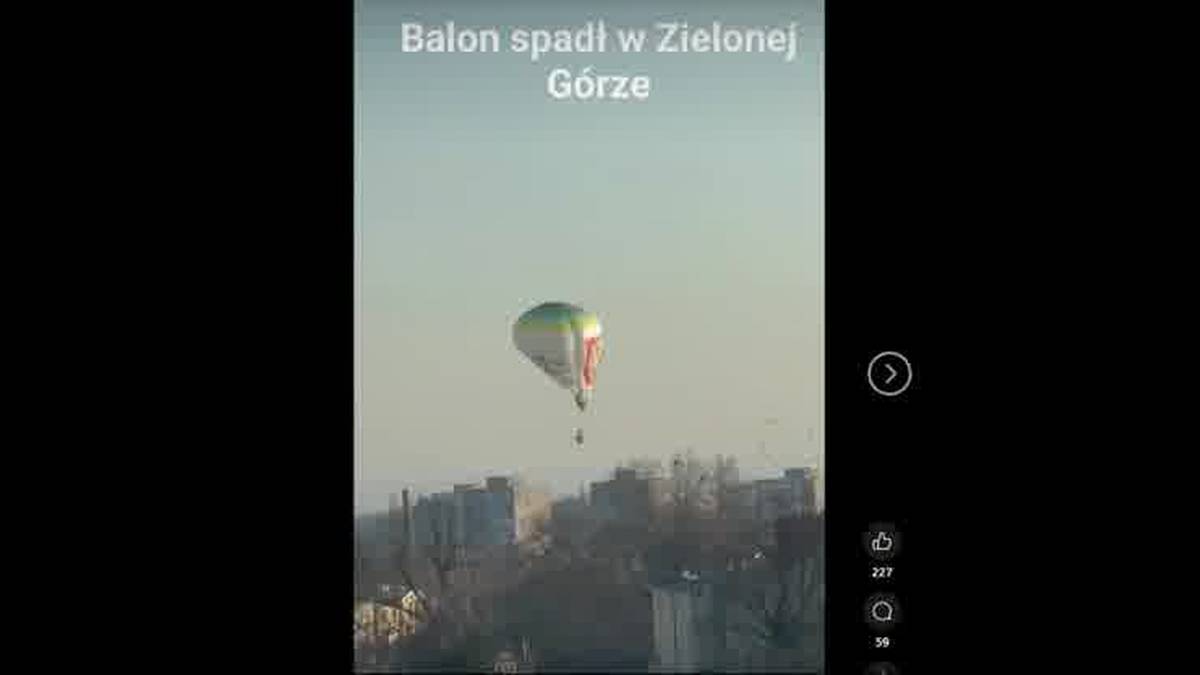 Balon o kolorze białym z zielonymi i czerwonymi pasami zawieszony na lince, opada w kierunku miasta.