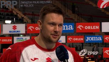 Portret Kamila Syprzaka, polskiego piłkarza ręcznego, podczas wywiadu. Ma na sobie biało-czerwoną koszulkę reprezentacji Polski. W tle widoczne są banery reklamowe sponsorów, w tym Suzuki, Orlen i Joma.