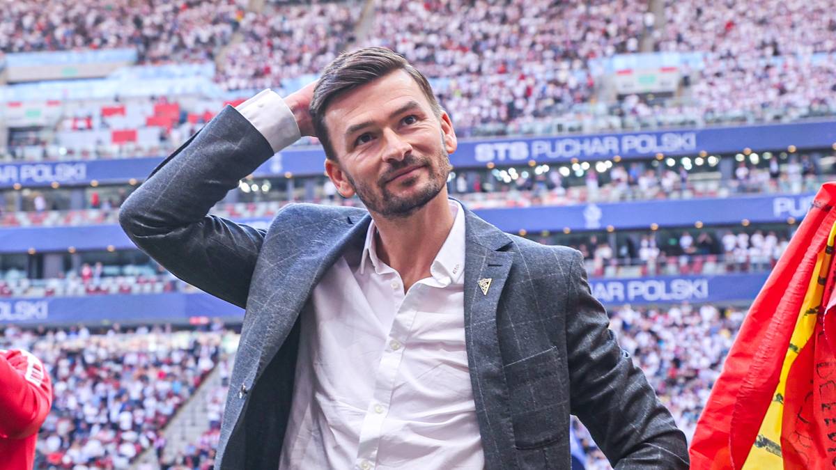 Trzech mężczyzn na stadionie, jeden w garniturze drapiący się po głowie, drugi z flagą na ramionach, trzeci klaszczący.