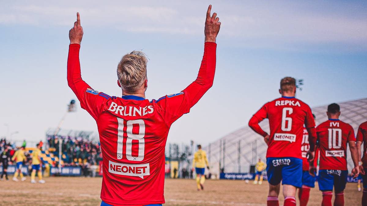 Piłkarz w czerwonej koszulce z numerem 18 na plecach, klęczy na boisku z rękami uniesionymi w g&oacute;rę w geście radości.