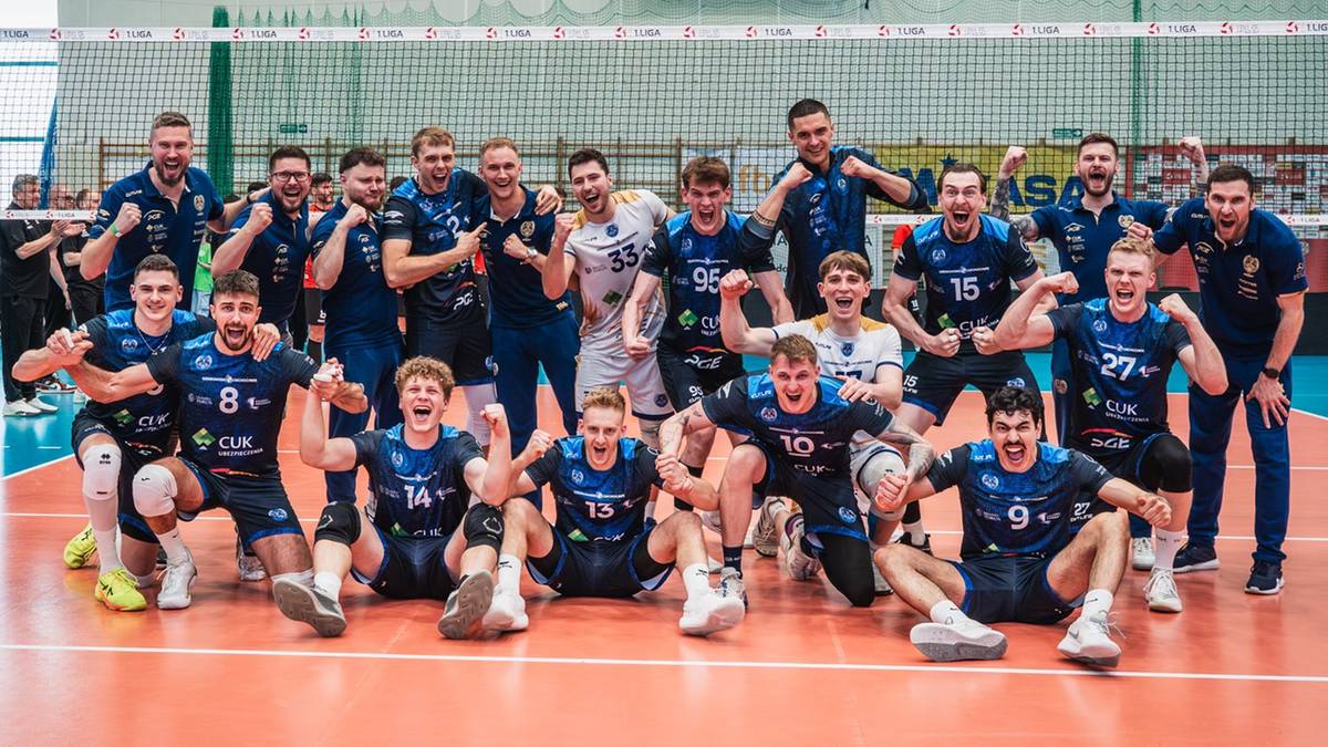 Grupa siatkarzy w niebieskich strojach sportowych, siedzących i stojących, pozuje do zdjęcia na boisku siatkarskim.