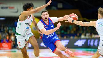 Gracz w niebieskim stroju z numerem 11 próbuje rzucić piłkę do kosza, podczas gdy dwaj przeciwnicy próbują go zablokować.