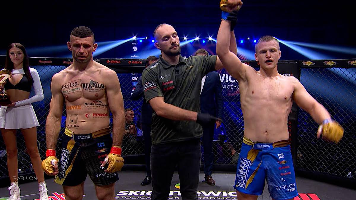 Sędzia podnosi rękę zwycięzcy walki MMA w klatce, obok stoi drugi zawodnik i ring girl.