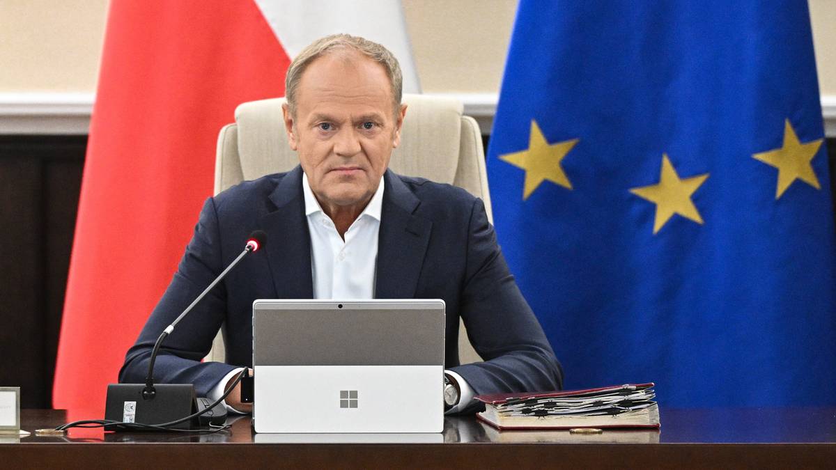 Premier Donald Tusk siedzi przy stole, przed nim stoi laptop i mikrofon. W tle widnieją flagi Polski i Unii Europejskiej.
