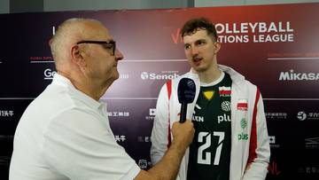 Michał Gierżot w białej kurtce z zielonymi elementami, udziela wywiadu, trzymając mikrofon. Dziennikarz z okulary w białej koszulce stoi przed nim. W tle logo "VOLLEYBALL NATIONS LEAGUE".