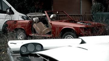 Czerwony zabytkowy samoch&oacute;d kabriolet w stanie rozkładu, stojący na zewnątrz wśr&oacute;d zarośli.