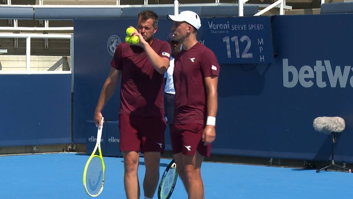 Dwóch mężczyzn w czerwonych strojach tenisowych stojących na korcie. Jeden z nich trzyma trzy piłki tenisowe w dłoni i jedną bierze do ust. Drugi mężczyzna ma na głowie białą czapkę.