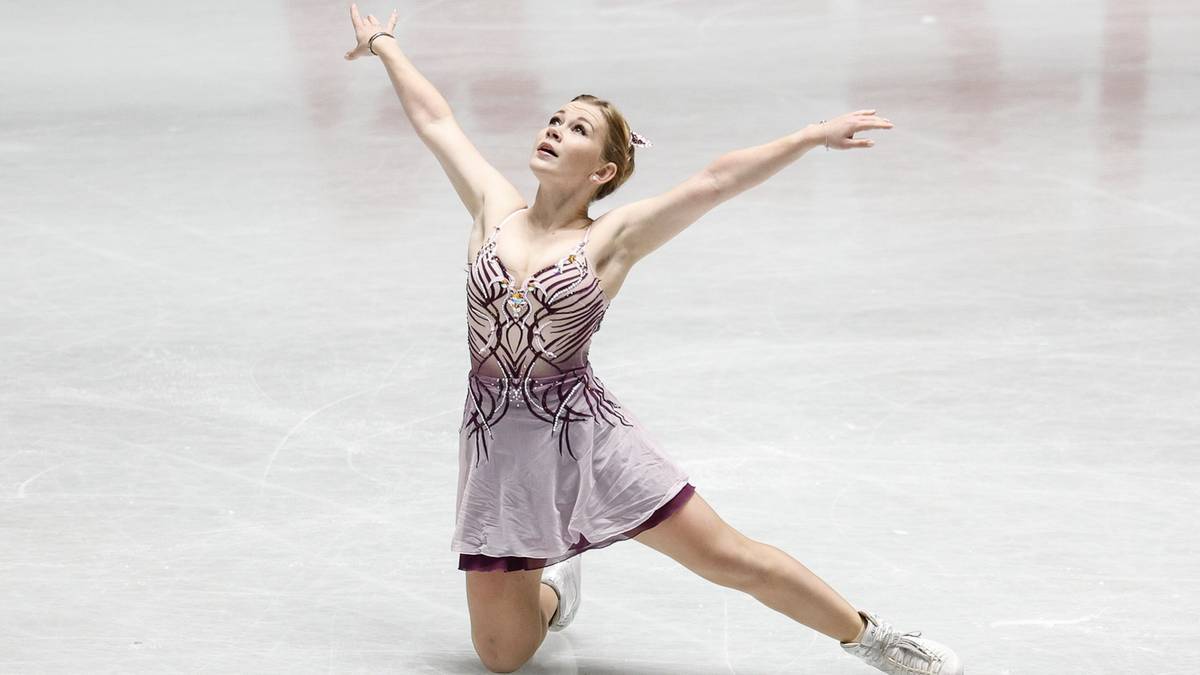 Łyżwiarka figurowa w fioletowej sukience z ozdobnymi wzorami na lodowisku, z uniesionymi ramionami i nogą w wykroku.