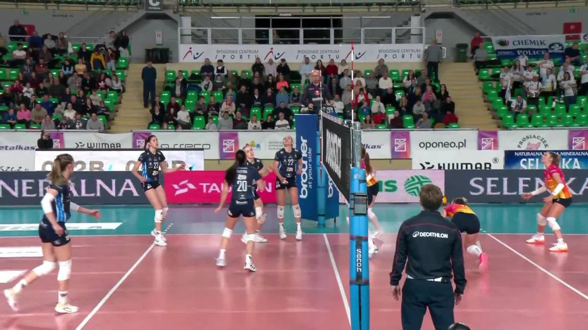 Siatkarki grające w meczu halowym, jedna zawodniczka w białym stroju biegnie do przodu, inne w ciemniejszych strojach po drugiej stronie siatki, w tle widzowie na trybunach.