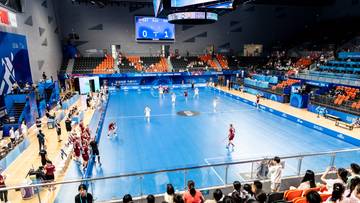 Drużyny unihokeja rozgrywają mecz na niebieskim boisku w hali sportowej, otoczonej trybunami pełnymi widzów. Widoczne są wyniki na tablicy.