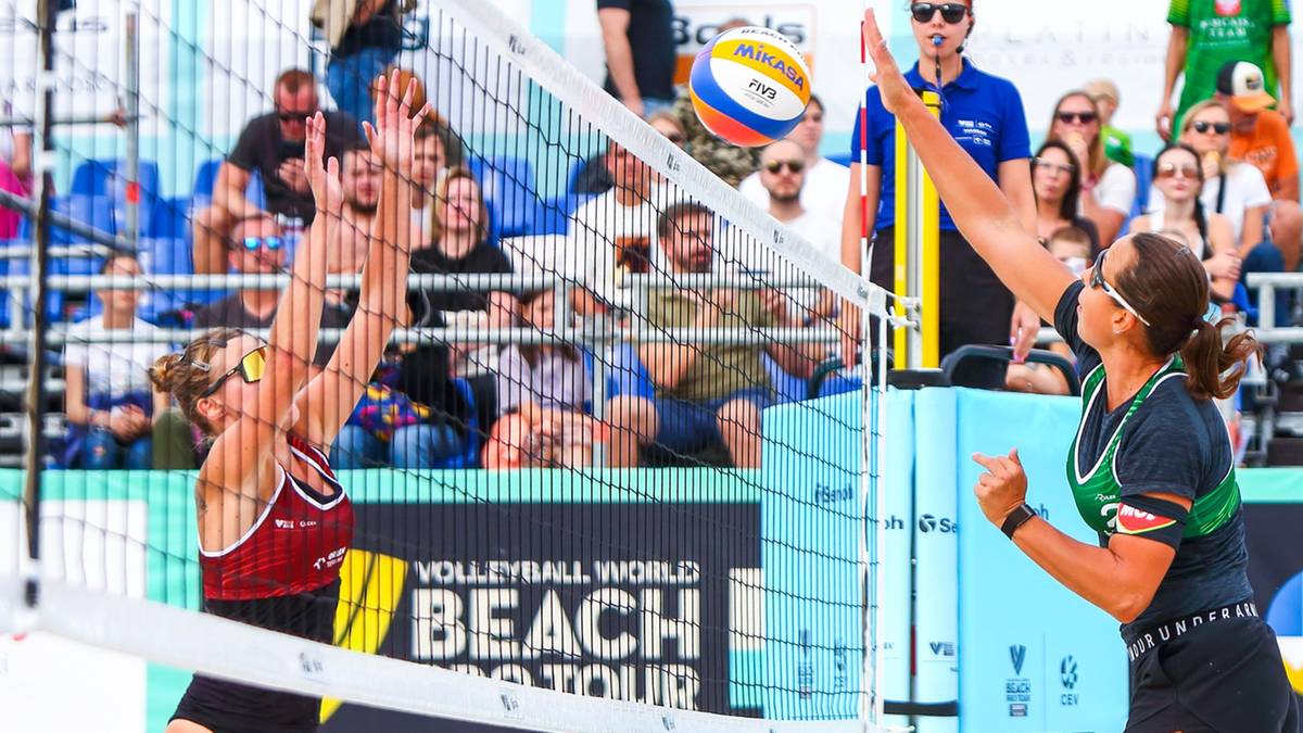 Dwie siatkarki w akcji, jedna z nich szykuje się do zagrywki, druga broni się przy siatce.