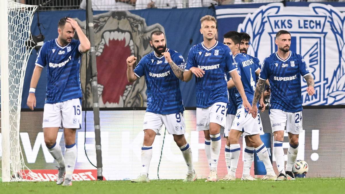 Sześciu piłkarzy Lecha Poznań w niebieskich koszulkach z białymi numerami i herbem klubu, stojący na boisku piłkarskim. Jeden z piłkarzy trzyma się za czoło, inni patrzą przed siebie lub na siebie. Za nimi widoczna jest trybuna i fragmenty stadionu.