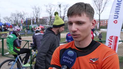 Mężczyzna w pomarańczowej koszulce z logo zespołu, wypowiadający się do mikrofonu z logo Polsat Sport. W tle inni sportowcy i rowery.