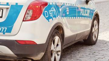 Tylna część białego samochodu policyjnego z niebieskimi pasami i napisem "POLICJA"