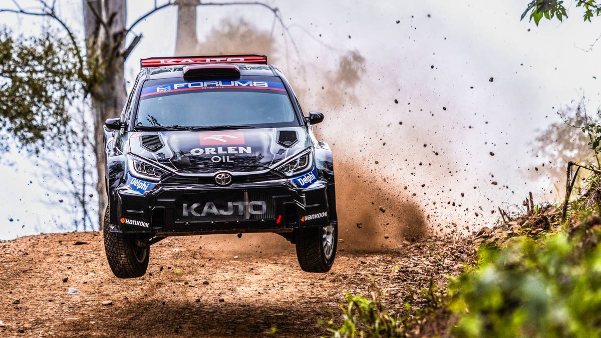 Samoch&oacute;d rajdowy z numerem 1 i napisem "KAJTO" na masce, w trakcie pokonywania zakrętu na piaszczystej trasie, unosząc tumany kurzu.