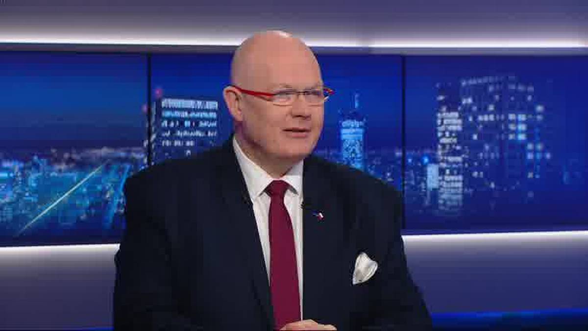 Mężczyzna w okularach i krawacie siedzi na tle wyświetlaczy z nocnym widokiem miasta. Ma ogoloną głowę i nosi ciemny garnitur.