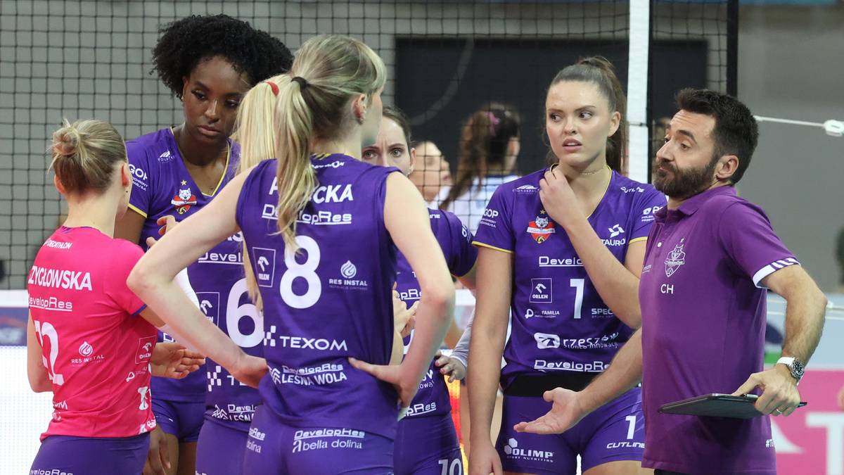 Grupa siatkarek w fioletowych strojach sportowych, z numerami na plecach, słuchająca wskazówek trenera.