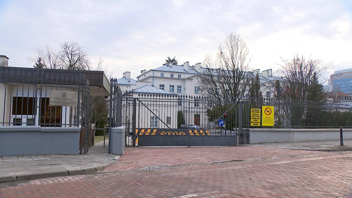 Brama wjazdowa do budynku z widocznym placem i drzewami w tle.