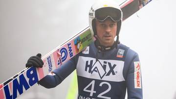 Skoczek narciarski w kasku i goglach, trzymający narty na ramieniu. Ma na sobie niebiesko-biały kombinezon z numerem 42.