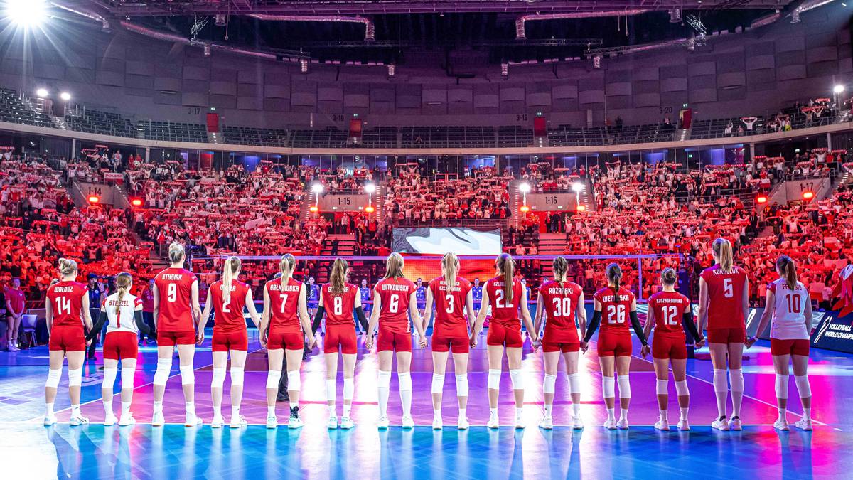 Drużyna siatkarek w czerwonych strojach pozuje na boisku przed liczną publicznością na trybunach.