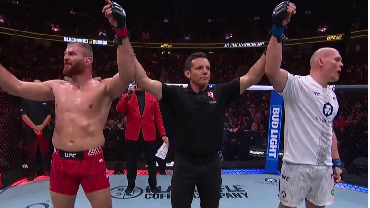 Sędzia unosi ręce dw&oacute;ch zawodnik&oacute;w MMA, Jana Błachowicza i Bogdana Guskova, po walce. Błachowicz stoi po lewej stronie, Guskov po prawej.