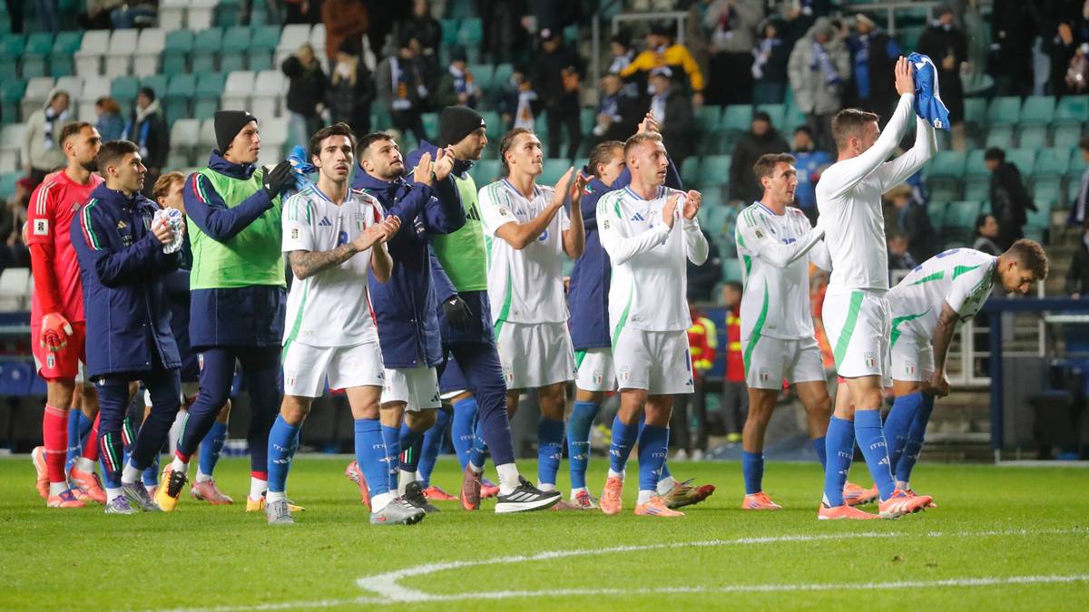 Drużyna piłkarska w białych strojach i błękitnych elementach świętuje na zielonej murawie stadionu, pozdrawiając kibiców na trybunach.
