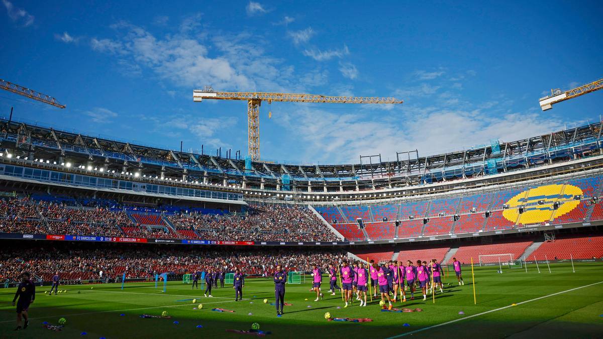 Stadion piłkarski Camp Nou z kibicami na trybunach i drużyną piłkarską trenującą na murawie, widoczne żurawie budowlane.