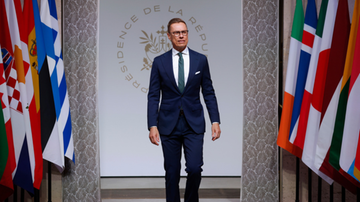 Prezydent Finlandii Alexander Stubb idzie w stronę kamery, otoczony flagami państwowymi, przed napisem &bdquo;PRESIDENCE DE LA REPUBLIQUE&rdquo;.