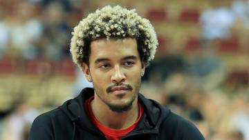 Portret mężczyzny z afro w blond włosy i brodą, ubranego w czarną bluzę z kapturem na czerwonym tle.