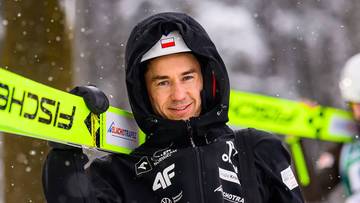 Polski skoczek narciarski z nartami na ramieniu, ubrany w zimową kurtkę i kask z polską flagą.