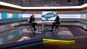 Dw&oacute;ch mężczyzn w garniturach rozmawia przy okrągłym stole w nowocześnie urządzonym studio telewizyjnym.