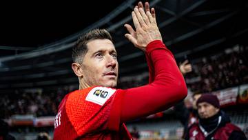 Robert Lewandowski w czerwonej koszulce z numerem i herbem klubu, klaszcze z uniesionymi dłońmi, na tle trybun stadionu.