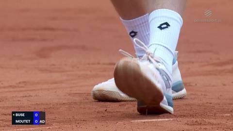 Zbliżenie na nogi tenisisty podczas meczu, widoczne buty sportowe i białe skarpetki na korcie ziemnym. W dolnym lewym rogu ekranu widoczny jest wynik meczu: Buse 0, Moutet 0.