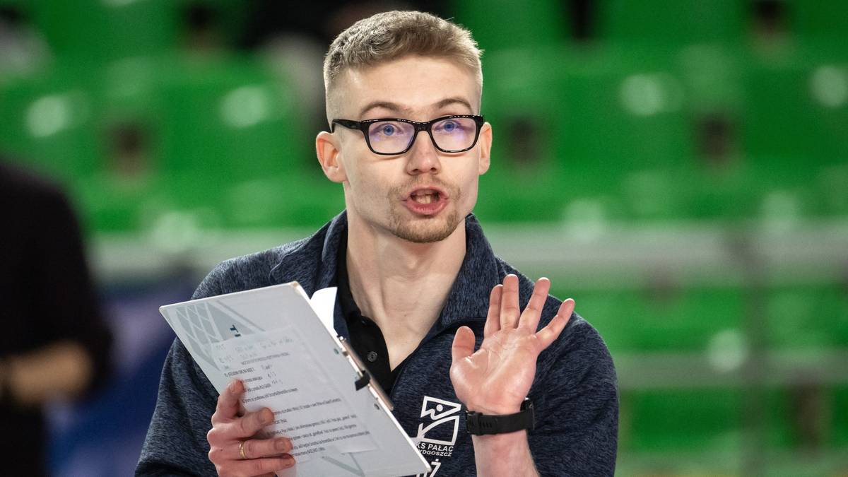 Młody mężczyzna w okularach trzyma dokument i gestykuluje podczas spotkania siatkarskiego.