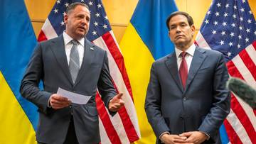 Dwóch mężczyzn w garniturach, jeden z nich trzyma dokument, stoi przed flagami USA i Ukrainy.