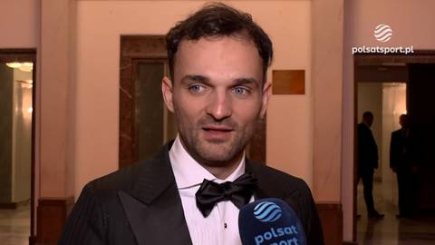 Bartosz Zmarzlik w garniturze i muszce, patrzący na wprost i trzymający mikrofon.