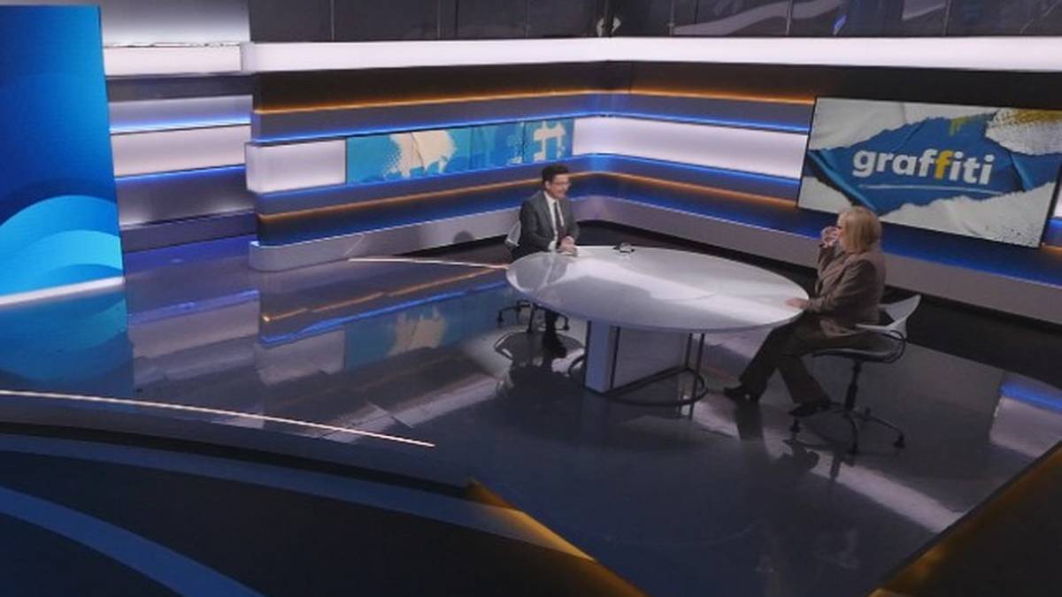 Dwie osoby siedzą naprzeciwko siebie w studiu telewizyjnym przy owalnym stole. W tle widoczne są ekrany z grafiką i napisami.