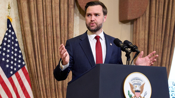 Mężczyzna w garniturze z krawatem przemawia za m&oacute;wnicą, obok niego flaga Stan&oacute;w Zjednoczonych.