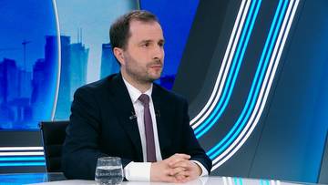 Mężczyzna w garniturze siedzi przy stole w studiu telewizyjnym.