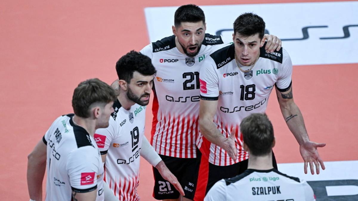 Pięciu siatkarzy w białych koszulkach z czerwonymi pasami, rozmawiających ze sobą na boisku siatkarskim.