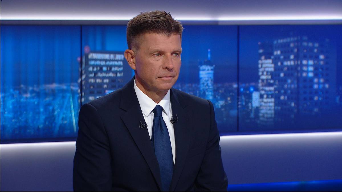 Mężczyzna w ciemnym garniturze i białej koszuli z granatowym krawatem siedzi przed ekranem z widokiem na nocne miasto.