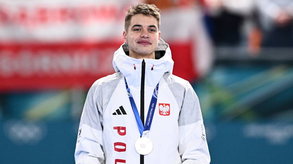 Polski sportowiec Semirunnij w białej kurtce z napisem "POLS" i z medalem na szyi, trzymający bukiet kwiat&oacute;w na podium.